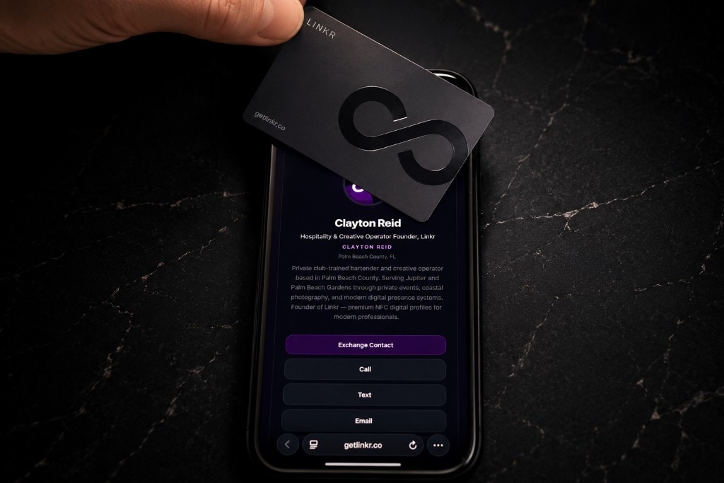 Hand holding LINKR matte black card above iPhone showing Clayton Reid profile on black marble — Exchange Contact